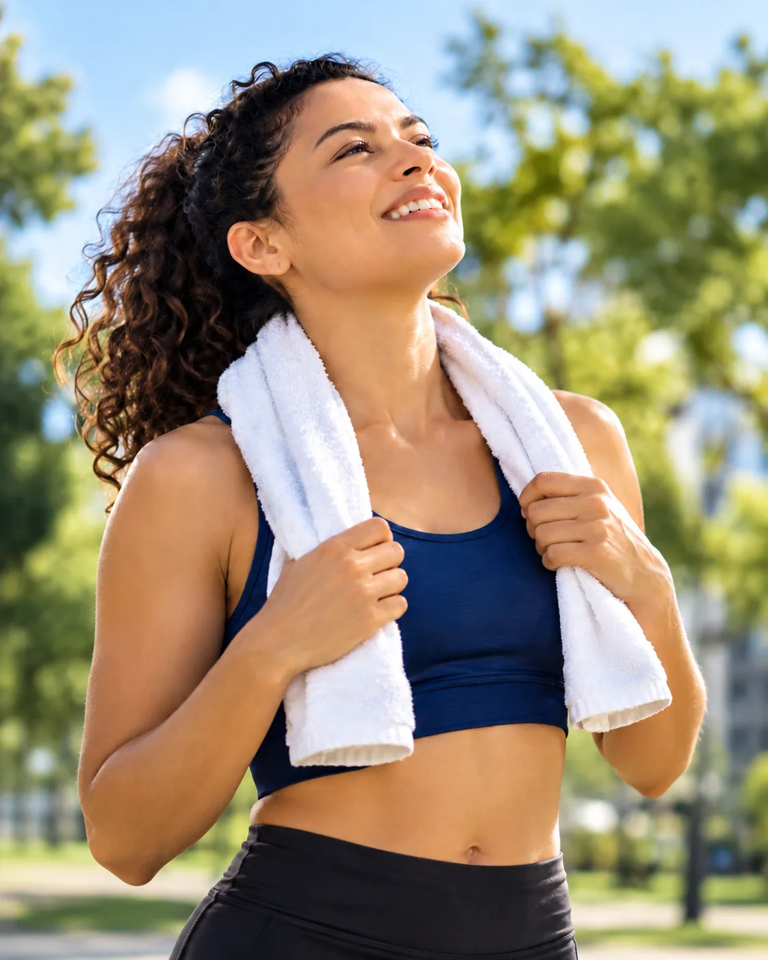 Mujer con ropa deportiva y una toalla al cuello sonríe mirando al cielo, en un parque